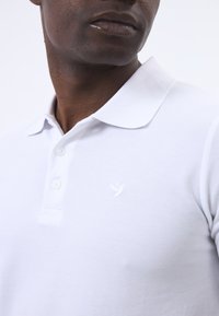 Bílá polo košile vyrobená z texturované tkaniny, s tříknoflíkovou légou a malým vyšitým logem ptáčka v bílé barvě na hrudi.
