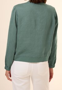 Chaqueta de lino verde holgada con cuello redondo, mangas fruncidas y dobladillo corto, combinada con pantalones blancos.