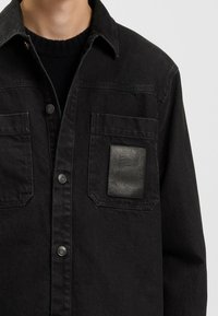 Zwarte denim shirt met een knoopsluiting aan de voorkant en twee borstzakken. Voorzien van een gestructureerde leren patch op de linkerzak.