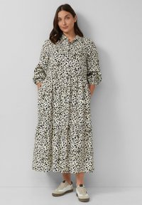 Un lungo abito a strati presenta uno sfondo crema con un motivo a stampa animale nera. L'abito ha una chiusura con bottoni sul davanti e tasche laterali.