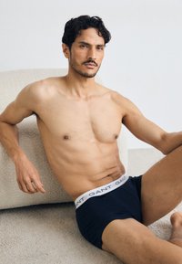 Svart boxertruser med hvit elastisk linning som har "GANT"-logo. Modellen sitter ned og viser fram en muskuløs overkropp og jevn hudstruktur.