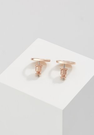 Paire de boutons de manchette en or rose avec des détails cylindriques et arrondis, posée sur un piédestal cubique blanc sur un fond gris clair.
