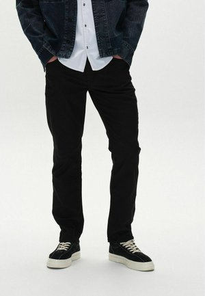  sorte bukser med lige pasform, parret med en hvid skjorte med knapper og en mørk denimjakke. Sorte sneakers med hvide såler fuldender looket.