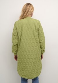 Kvinde med langt blond hår har en lys grøn quiltet lang jakke og blå jeans på, stående med ryggen til foran en ensfarvet hvid baggrund.