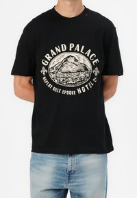 Schwarzes Baumwoll-T-Shirt mit zentralem Grafikmotiv eines Berges und dem cremefarbenen Schriftzug "GRAND PALACE REPLAY BELLE EPOQUE HOTEL". Kurze Ärmel, lockere Passform.
