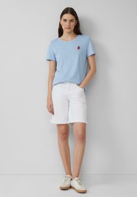 lichtblauwe katoenen t-shirt met een kleine rode aardbeienborduursel, gecombineerd met witte shorts met omgeplooide zoom en beige sneakers op een neutrale achtergrond.