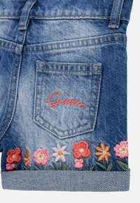 Plave traperice kratke hlače s cvjetnim vezom na rubu, u nijansama ružičaste, narančaste i zelene. Imaju stražnji džep s "Guess" logom.