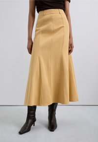 Femme portant une longue jupe jaune moutarde taille haute avec des coutures verticales, associée à des bottes noires à talons aiguilles sur un sol gris.