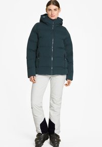 Giacca piumino verde scuro con cappuccio e zip frontale, abbinata a pantaloni da sci grigio chiaro con accenti blu navy e stivali da sci neri.