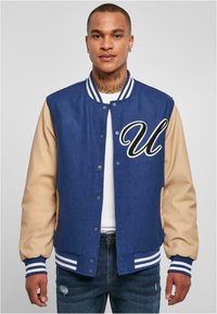 Jachetă varsity în nuanțe de navy și bej, cu un corp din lână, mâneci din piele, dungi albe la guler și manșete, având brodat pe piept un mare "U".