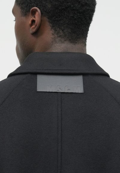 Vedere din spate a unei persoane cu păr scurt și creț, purtând o haină întunecată cu o patch rectangulară pe spatele superior care scrie "MSGM".
