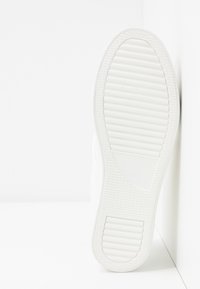 Suola bianca di sneaker con scanalature e motivi testurizzati, caratterizzata da bordi lisci e un design piatto, appoggiata su uno sfondo bianco.