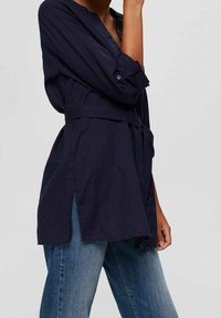 Chemise bleu marine ceinturée à manches longues, col à revers, fentes latérales et coupe décontractée. Confectionnée en tissu léger. Portée avec un jean bleu.