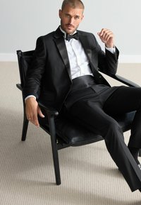 Schwarzer Smoking mit satinierter Revers, weißem Hemd und Fliege. Sitzt auf einem schwarzen Stuhl, im Hintergrund ein strukturierter Teppich, entspannte Pose.