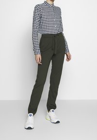 Person, die ein navyblaues und weißes karriertes Kurzarmhemd, dunkelgrüne, taillierte Hose und weiße Sneaker mit blauen Akzenten trägt.