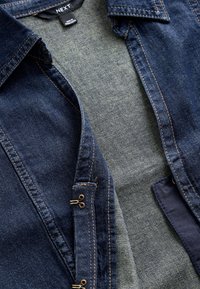 Veste en denim avec coutures marron, col et fermetures à crochets, montrant une doublure intérieure grise et une étiquette de marque "NEXT".