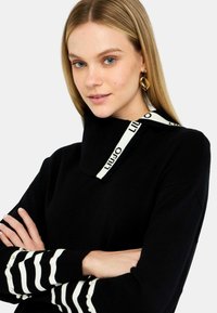 Maglione nero con colletto alto, caratterizzato da una fascia bianca con logo. Polsini a righe accentuati da piccole perle bianche. Texture morbida.
