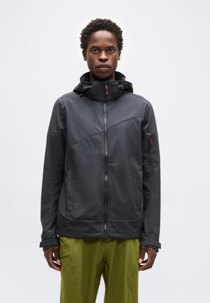 Nuori mies, jolla on musta vetoketjullinen huppari ja oliivinvihreät housut, seisoo vaaleaa yksiväristä taustaa vasten.