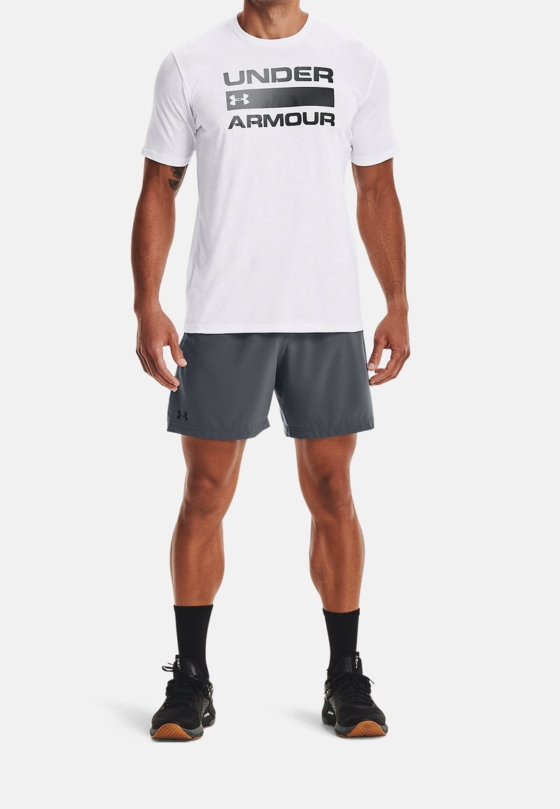 Vit t-shirt med korta ärmar och svart text "UNDER ARMOUR", kombinerad med mörkgrå shorts, svarta strumpor och svarta sportskor med orange detaljer.
