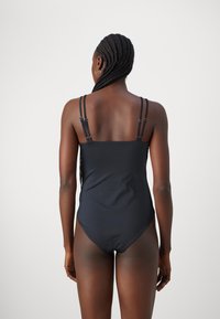 Et svart heldekkende badedrakt med en jevn tekstur, som har tynne justerbare stropper og en klassisk skjæring. Bakdesignet er enkelt og elegant.