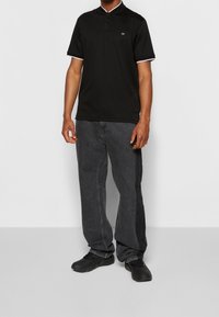 Polo noir à bordure blanche, manches courtes, avec logo CK, associé à un jean gris foncé ample et des chaussures noires.