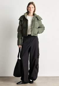 Veste doudoune verte avec un design court, des boutons-pression et un col montant ; associée à un pantalon cargo noir ample et un sac fourre-tout noir.