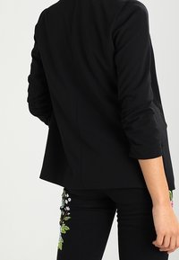 Blazer noir sur mesure avec une texture lisse, une coupe ajustée et des manches longues. Présente une broderie florale sur un jean foncé.