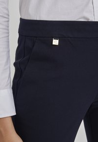 Granatowe, dopasowane spodnie o gładkiej fakturze, z akcentem w postaci złotego kwadratowego logo na pasku i bocznych kieszeniach.