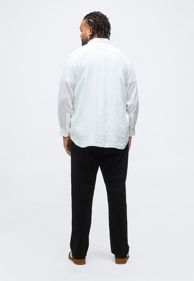 Άνδρας με πλεγμένα μαλλιά στραμμένος προς τα πίσω, φορώντας λευκό πουκάμισο με μακριά μανίκια, μαύρο παντελόνι και μαύρα παπούτσια με καφέ σόλες, μπροστά σε ένα απλό φόντο.