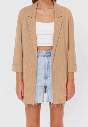 Femme portant un blazer beige à fines rayures sur un crop top blanc et un short en denim bleu clair taille haute aux bords effilochés.