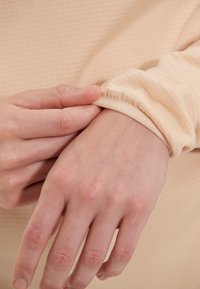 Langærmet top med et tekstureret lysebeige stof. Elastiske cuff med let rynkning, hånd der blidt trækker i ærmet. Glat hudtone synlig.