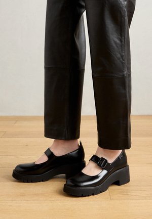 Persoon draagt zwarte leren broek en zwarte chunky Mary Jane platformschoenen, staand op een houten vloer.