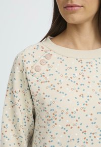 Felpa beige con motivi astratti in blu, arancione e bianco. Presenta tre bottoni decorativi sulla spalla sinistra. Texture morbida.