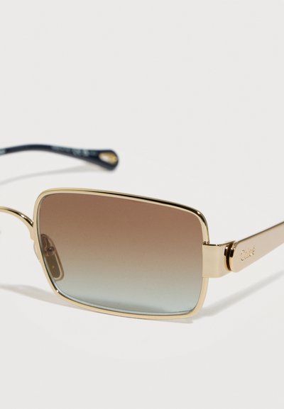 Ορθογώνια γυαλιά ηλίου με χρυστό μεταλλικό σκελετό και φακούς με διαβάθμιση από καφέ σε μπλε απόχρωση, εμφανίζονται σε κοντινό πλάνο σε λευκό φόντο.