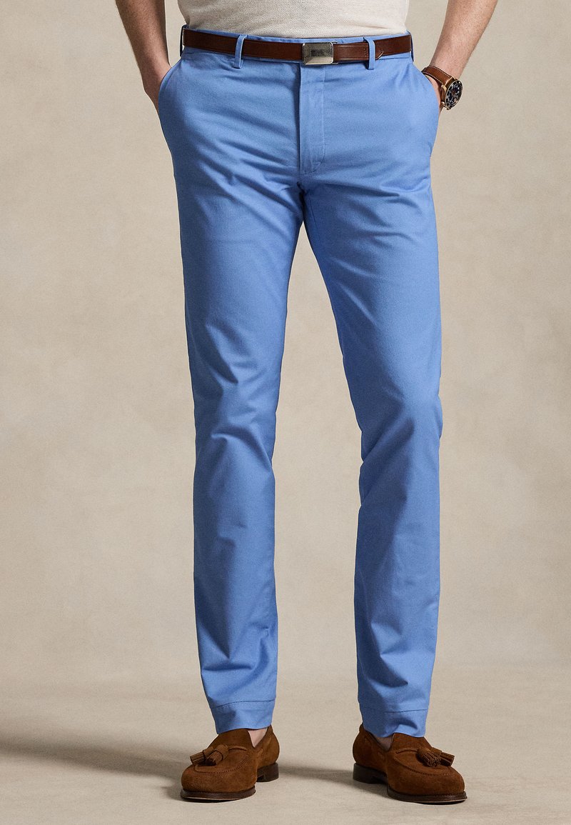 Άνδρας που φοράει ανοιχτό μπλε παντελόνι slim-fit, καφέ δερμάτινη ζώνη, καφέ σουέντ μοκασίνια με φούντες και ρολόι χειρός, στέκεται με τα χέρια στις τσέπες.