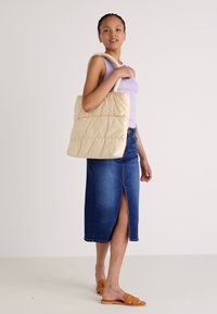 Sac fourre-tout beige matelassé avec des poignées arrondies, texture douce et motif subtil. Le modèle porte un haut lavande et une jupe en denim.