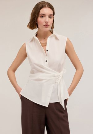 Witte mouwloze blouse met een kraag, knoopsluiting en een gestrikt taille-detail. Gladde katoenen stof, gecombineerd met donkerbruine wijde broekspijpen.