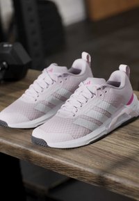Ζευγάρι ανοιχτό ροζ παπούτσια Adidas για τρέξιμο με λευκές ρίγες σε ξύλινη επιφάνεια, με θολά γυμναστήρια στον φόντο.