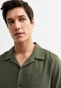 Chemise olive verte à manches courtes avec un col, dotée d'une poche poitrine, d'une fermeture à boutons et d'un tissu texturé.