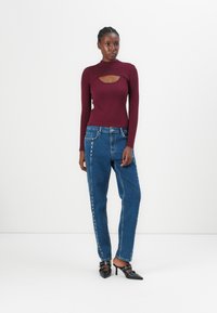 Bordeaux langearmige top met een uitsnijding, gecombineerd met blauwe high-waisted jeans met zijkantstuds en zwarte hakken met accenten.