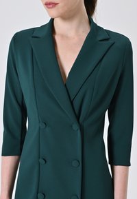 Dunkelgrüner, doppelt geknöpfter Blazer mit Reverskragen, Dreiviertelärmeln und mit Stoff überzogenen Knöpfen, der eine glatte Textur aufweist.