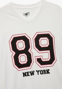 Hvid sports t-shirt med fed sort og rød grafisk tekst "89" og "NEW YORK," med v-halsdesign og tekstureret stof.