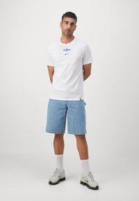 Weiße Baumwoll-T-Shirt mit blauem Grafiklogo, kombiniert mit hellblauen Jeansshorts und hellgrauen Sneakers mit schwarzen Akzenten.