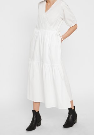 Kobieta w białej, midi sukience kopertowej z bufkami na rękawach, ubrana w czarne botki za kostkę, stojąca z jedną ręką w kieszeni.