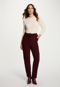 Mujer de pie con la mano en el bolsillo, llevando un suéter de canalé crema, pantalones burdeos de cintura alta con cinturón y botas de tobillo burdeos.
