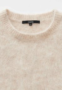 Pull beige clair en matière douce et duveteuse. Il présente un col rond classique et une texture de tricot simple et lisse. Étiquette de taille 36 visible.