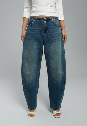 Personne portant un jean en denim bleu ample, un haut blanc à manches longues et des sandales blanches à talons ouverts, debout sur un fond gris.