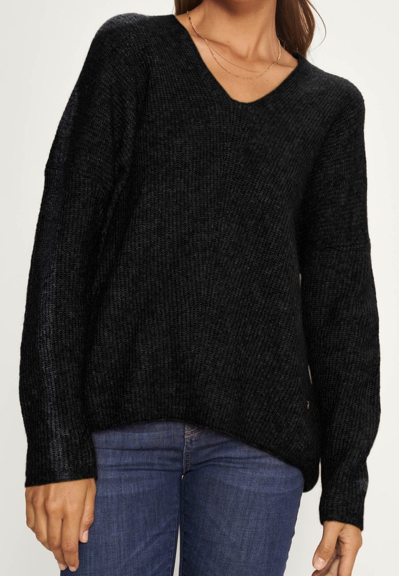 Femme portant un pull en maille noire à col en V et un jean bleu, avec un collier délicat en or visible autour de son cou.