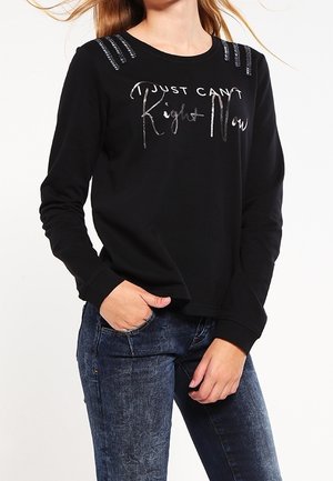 Kvinde iført sort langærmet skjorte med sølvtekst "Just Can't Right Now" og dekorative striber på skuldrene, kombineret med blå jeans.