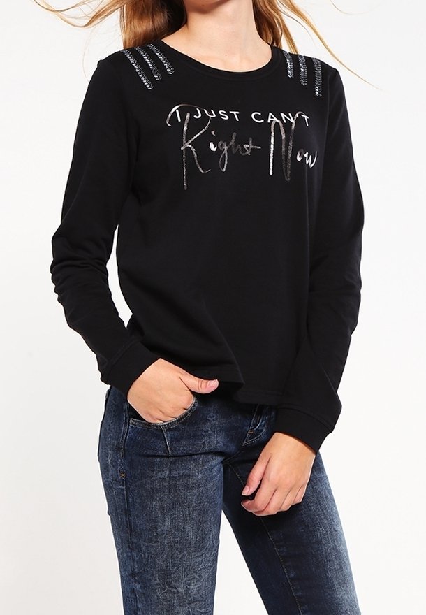 Femme portant un haut noir à manches longues avec le texte argenté « Just Can't Right Now » et des bandes décoratives sur les épaules, associé à un jean bleu.